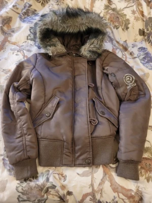 Supersnygg jacka i brun med pälsluva - Supersnygg jacka i brun med pälsluva. Mycket fint skick bortsett från att en knapp saknas och att de andra sitter lite löst och litet skraphål på ena armen, men inget som riktigt märks när man bär den (se bilder för egen bedömning). Annars fint skick. Vid frågor är det bara att skriva:)