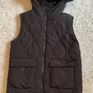 Svart dunväst med huva - Snygg svart dunväst med quiltat mönster och huva. Västen har dragkedja framtill och två stora fickor med lock. Perfekt för lager-på-lager och ger en cool streetwear-vibe. Materialet är mjukt och puffigt för extra värme. Strl 130, använd få gånger. 