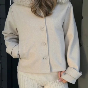 Beige ullkappa med fuskpäls - Stilren beige kappa i ullmix med stor krage i fuskpäls. Kappan har raka ärmar, knäppning med fyra knappar framtill och två sidofickor. Perfekt för kyliga dagar och ger en lyxig känsla till din outfit. Använd 1-2 gånger❤️❤️
