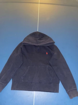 Mörkblå hoodie från Polo Ralph Lauren - Snygg mörkblå hoodie från Polo Ralph Lauren med klassisk röd logga broderad på bröstet. Hoodien har en stor huva och en praktisk magficka framtill. Perfekt för en avslappnad och stilren look.