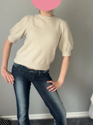 Krämvit puffärm stickad tröja - Säljer en krämvit stickad tröja med puffärmar och ribbade muddar. Tröjan har rund halsringning och kortare ärmar som ger en trendig siluett. Perfekt att styla med jeans eller kjol för en cool look.