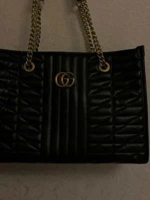 Svart handväska  - Snygg svart handväska med guldfärgade kedjehandtag. Väskan har ett rymligt fack med ljus insida och innerficka med dragkedja. 