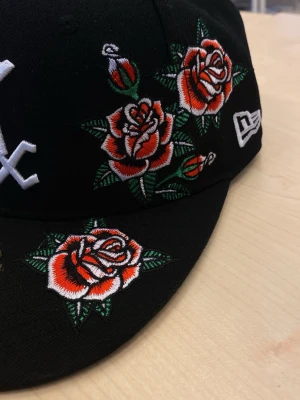 Svart New Era keps med rosbrodyr - Snygg svart keps från New Era med broderade röda och vita rosor samt gröna blad. Kepsen har en klassisk form, platt skärm och vita detaljer, inklusive ett broderat emblem på sidan. Materialet är bomull och modellen är fitted.  7 3/8 storlek
