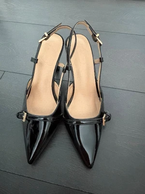 Svarta klackar med guld detalj  - Säljer ett par svarta pumps i lack med spetsig tå och smal klack. Skorna har öppen häl med justerbara remmar och guldfärgade spännen. Storlek 34 men passar till 35 också! Säljer eftersom fick 2 stycken exakt likadana i present. 