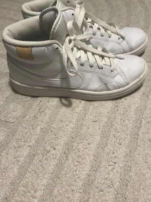 Vita Nike high top sneakers - Säljer ett par klassiska vita high top sneakers från Nike. Skorna har en stilren design med vita skosnören, diskret swoosh på sidan och en liten beige detalj vid hälen. Tillverkade i läder med vadderad insida för extra komfort. Perfekt till vardag eller kanske utspring på studenten! Använda några gånger.