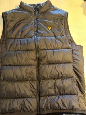 Mörkgrå dunväst från Lyle & Scott - Snygg mörkgrå dunväst från Lyle & Scott med gul logga på bröstet. Västen har hög krage, dragkedja framtill och breda quiltade paneler. Perfekt för lager på lager och håller dig varm under kyliga dagar. Materialet är glansigt och lätt.