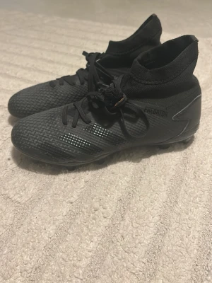 Svarta Adidas Predator fotbollsskor - Säljer ett par svarta Adidas Predator fotbollsskor med hög strumpkrage och snörning. Skorna har texturerad ovandel för bättre bollkontroll och FG-dobbar för spel på gräs. Tillverkade i syntetmaterial och designade för att ge bra grepp och stabilitet på planen. Inte använda alls, säljs inte längre men ordinare pris var 500kr. Unisex