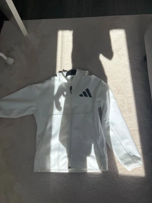 Vit hoodie från Adidas med dragkedja - Snygg vit hoodie från Adidas med dragkedja framtill och svart Adidas-logga på bröstet. Hoodien har huva och praktiska fickor, varav en med dragkedja. Tillverkad i mjukt material som känns skönt mot huden. Perfekt för en sportig och avslappnad stil.
