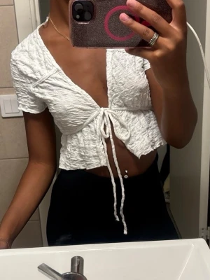Vit croppad topp med snörning - Trendig vit croppad topp med korta ärmar och snygg snörning framtill. Toppen har en skrynklig textur och v-ringad halsringning som ger en cool och avslappnad look. Perfekt att matcha med höga byxor eller kjol.