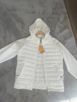 Vit vadderad vindjacka med huva - Snygg vit vindjacka med quiltad framsida och huva. Jackan har dragkedja framtill och två fickor med dragkedja. Tillverkad i ett mjukt material med släta ärmar och ribbad nederkant. Perfekt för dig som vill ha en stilren och sportig look.