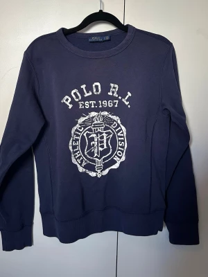 Mörkblå sweatshirt Polo Ralph Lauren - Mörkblå sweatshirt från Polo Ralph Lauren med vit tryckt logga och text 'POLO R.L. EST. 1967' på bröstet. Rund halsringning och ribbade muddar vid ärmslut och nederkant. Perfekt för en avslappnad och sportig stil.