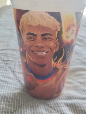 McDonald's plastmugg med fotbollstryck - Cool plastmugg från McDonald's med färgglatt tryck av en tecknad fotbollsspelare, eldflammor och fotbollstema. Muggen har vita kanter och motiv i rött, blått, gult och svart. Perfekt för dig som gillar sportiga samlarprylar.