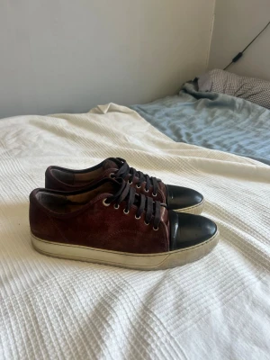 Vinröda sneakers med svart toebox från Lanvin - Snygga vinröda sneakers i mocka med svart tåparti i läder och svarta skosnören. Skorna har vit sula och klassisk låg profil. Perfekta för dig som vill ha en stilren men ändå unik look. Om du vill hitta de perfekta sommarskorna, kolla in dessa! Priset kan förhandlas