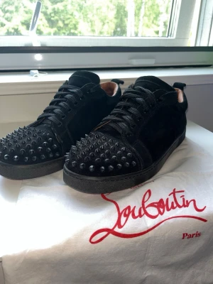 Svarta Louboutin sneakers med nitar - Säljer ett par svarta sneakers från Christian Louboutin med coola svarta nitar över tån och klassisk röd sula. Skorna är låga och har snörning samt är gjorda i mocka och läder. Perfekta för dig som vill sticka ut med en edgy stil.
