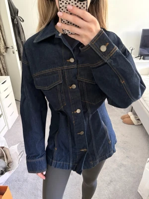 Mörkblå figursydd jeansjacka - Snygg mörkblå jeansjacka (går även att ha som jeansskjorta) i storlek XS med markerad midja och klassiska bronsfärgade knappar. Jackan har två bröstfickor med lock och kontrastsömmar. Aldrig använd med prislapp på! Finns ett band innanför som man kan fästa hur man vill beroende på hur figurnära eller lös man vill att den ska vara. 
