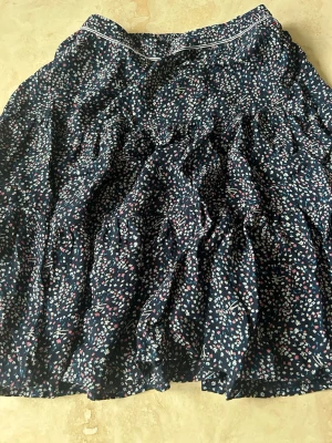 Blommig mörkblå volangkjol - Söt mörkblå kjol med småblommigt mönster i vitt, rosa och blått. Kjolen har volanger och en tunn vit kant upptill som detalj. Den har en bra passform vid midjan.