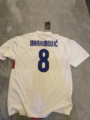 Inter Milan Ibrahimovic matchtröja - Säljer en vit Inter Milan matchtröja från Nike med röd korsdesign, klubbmärke och Pirelli-logga på bröstet. På ryggen står Ibrahimovic och nummer 8 i blått. Perfekt för dig som älskar fotboll och vill ha en ikonisk supportertröja.
