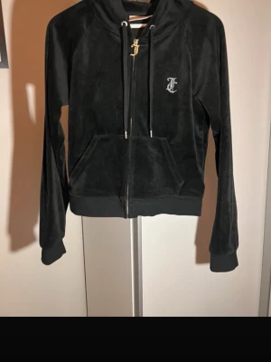 Svart velour hoodie från Juicy Couture - Snygg svart hoodie i mjuk velour från Juicy Couture. Dragkedja framtill, fickor och huva med snören. Juicy-logga i strass på ryggen och ett litet J på bröstet. Perfekt för en chill och trendig look.