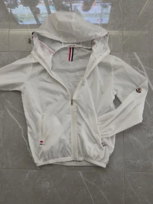 Vit vindjacka från Moncler - Snygg vit vindjacka från Moncler med huva och dragkedja framtill. Jackan har tunnt, lätt material och detaljer i rött, vitt och blått på huva och dragkedja. Moncler-logga på ärmen och justerbar dragsko vid huvan. Perfekt för blåsiga dagar.