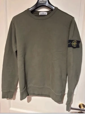 Olivgrön sweatshirt från Stone Island - Säljer en olivgrön sweatshirt från Stone Island med klassisk rund hals och ribbade muddar. Tröjan har den ikoniska Stone Island-loggan på vänster ärm och är tillverkad i mjuk bomull. Perfekt för en avslappnad och stilren look.
