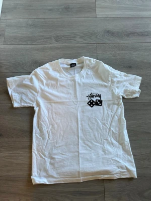 Vit Stüssy t-shirt med tärningsprint - Vit t-shirt från Stüssy med svart logga och två tärningar tryckta på bröstet. Klassisk rund hals och korta ärmar. Perfekt för dig som gillar streetwear och enkel stil.