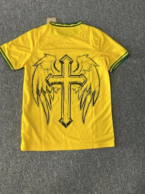 Gul Brasil tröja med Jesusmotiv - Säljer en gul Brasilien-tröja från Nike med gröna detaljer på krage och ärmslut. Framsidan har ett stort motiv av Jesus med törnekrona och Brasil-logga, baksidan har ett kors med vingar. Tillverkad i lätt och ventilerande material, perfekt för fotboll eller streetwear.