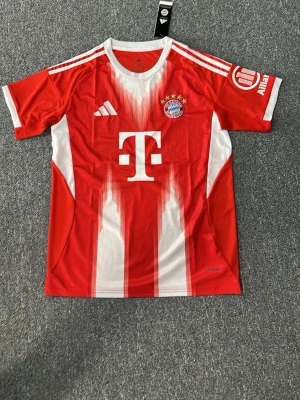 FC Bayern München matchtröja Adidas - Röd och vit matchtröja från Adidas för FC Bayern München. Tröjan har klubbmärke, Adidas-logga och sponsortryck på bröstet samt tryck på ryggen. Tillverkad i lätt och ventilerande material, perfekt för fotbollsträning eller match.