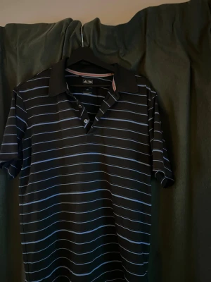 Golfpike från Adidas - Snygg svart pikétröja från Adidas med tunna vita och ljusblå ränder. Klassisk krage och knappslå framtill. Tillverkad i ett lätt och bekvämt material som passar perfekt till sport eller golf. Adidas-logga i nacken och diskreta detaljer vid kragen.