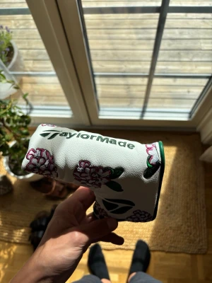 TaylorMade putter headcover (Masters edition) - Säljer ett (Masters edition) vitt putter headcover från TaylorMade med broderade rosa och gröna blommor. Skyddet har en klassisk form och är gjort i konstläder med gröna detaljer och TaylorMade-logga. Perfekt för dig som vill ha en snygg och unik look på golfbanan.