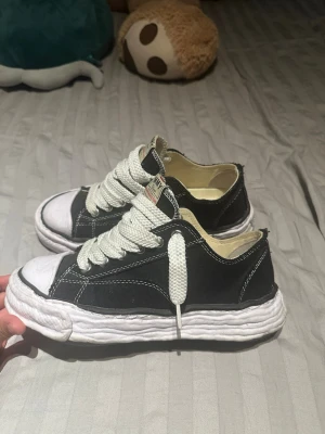 Svarta Maison Mihara låga sneakers - Svarta sneakers från Maison Mihara med vit tjock sula och breda vita skosnören. Skorna har en klassisk låg silhuett och kontrastsömmar i vitt. Ovandelen är i tyg och sulan har en chunky look. Perfekta för dig som gillar streetwear och vill sticka ut.