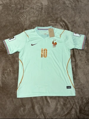 Frankrike fotbollströja Mbappé #10, S,M, L  - Snygg ljusgrön fotbollströja från Nike med Frankrikes landslagsmärke och nummer 10 i guld. Mbappé står på ryggen och tröjan har detaljer i orange och lila vid ärmslut. Perfekt för dig som älskar fotboll och vill stötta Frankrike.