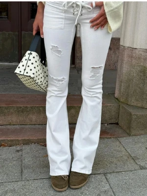 Vita bootcut jeans med snörning - Säljer ett par vita bootcut jeans med snörning i midjan och slitna detaljer på låren och knäna. Jeansen har hög midja och utsvängda ben, vilket ger en trendig siluett. Perfekta för dig som vill sticka ut med en cool och modern look.