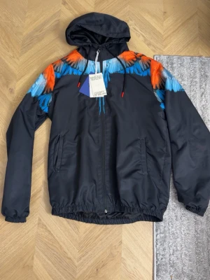 Svart vindjacka Marcelo Burlon - Svart vindjacka med huva från Marcelo Burlon. Jackan har en färgstark vingdesign i orange och blått över axlarna, dragkedja framtill och två sidofickor. Justerbar huva med svarta snören och röda detaljer. Perfekt för en cool streetwear-look. Den passar en stor M och L-xl. Använd ett par gånger.