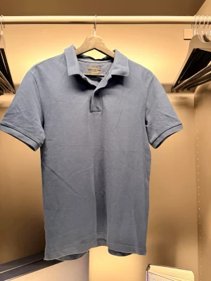 Massimo Dutti - Klassisk blå piké t-shirt med krage och korta ärmar. Tillverkad i mjuk bomull och har en enkel, stilren look utan synliga loggor eller mönster. Perfekt till jeans eller shorts för en avslappnad stil.