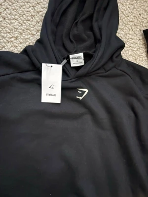 Svart hoodie från Gymshark - Svart hoodie från Gymshark med stor vit logga på ryggen och mindre logga på bröstet. Perfekt för gymmet eller chill. Mjuk och skön bomullsblandning. Passar även storlek S