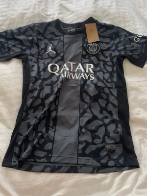 Svart PSG x Jordan matchtröja - Snygg svart och grå PSG matchtröja i samarbete med Jordan. Tröjan har camo-mönster, korta ärmar och tryck med Qatar Airways på bröstet. PSG-logga och Jumpman-logga på framsidan. Perfekt för fotboll eller streetwear.