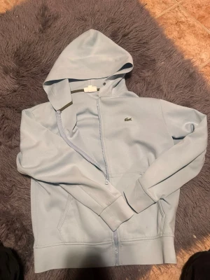 Ljusblå hoodie från Lacoste - Snygg ljusblå hoodie från Lacoste med dragkedja framtill och klassisk krokodil-logga på bröstet. Hoodien har huva med dragsko och två fickor framtill. Tillverkad i mjukt bomullsmaterial som känns skönt mot huden. Perfekt för en avslappnad och stilren look.