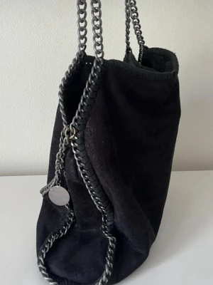 Svart väska i falabella-stil - Säljer en jättesnygg svart tote-väska i mockaimitation med silverfärgade kedjordetaljer längst kanterna. Superfin väska, skriv om frågor. Vill gärna få sålt snabbt, så kom med prisförslag