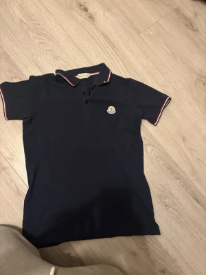 Marinblå pikétröja från Moncler - Snygg marinblå pikétröja från Moncler med klassisk krage och knappar framtill. Tröjan har röda och vita detaljer på krage och ärmslut samt Moncler-logga på bröstet. Perfekt för en clean och sportig look.