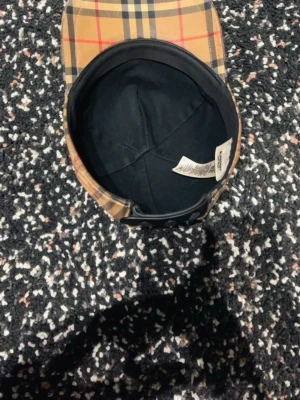 Burberry beige rutig keps - Säljer en klassisk Burberry keps i beige med det ikoniska rutmönstret i svart, vitt och rött. Kepsen har justerbar rem bak och är tillverkad i bomull. Perfekt accessoar för dig som vill ha en tidlös och trendig look.