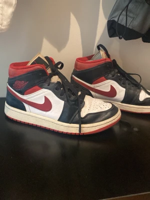 Air Jordan 1 Mid svart/röd/vit - Säljer ett par Air Jordan 1 Mid sneakers från Nike i svart, rött och vitt läder. Klassisk design med röd Swoosh, svart snörning och röd yttersula. Wings-logga på sidan och Air-märke på plösen. Perfekta för dig som gillar streetwear och ikoniska sneakers.
