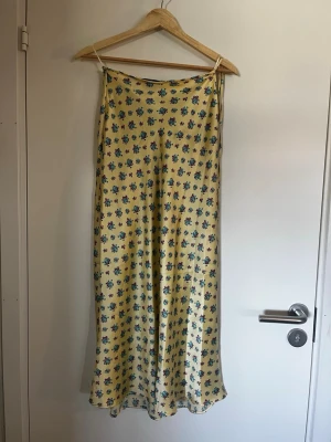 Gul blommig lång kjol - Säljer en söt gul lång kjol med små blommor i blått och rött. Kjolen har snörning på ena sidan och dragkedja på andra och ett mjukt, följsamt material som ger en avslappnad look. Perfekt för varma dagar och sommarens alla tillfällen.