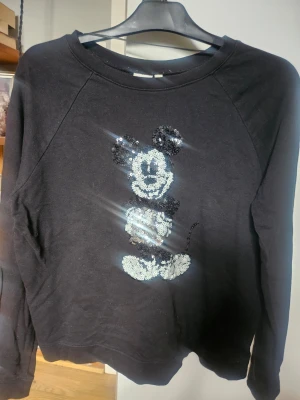 Svart sweatshirt med paljett-Musse Pigg  - Cool svart sweatshirt med Musse Pigg-motiv i svartvita paljetter på bröstet. Tröjan har rund halsringning och långa ärmar. Perfekt för dig som gillar Disney och vill ha något som sticker ut i garderoben.