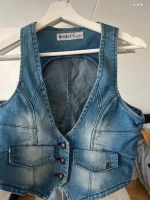 Jeansväst från Monica's - Snygg jeansväst i blå denim från Monica's med markerade sömmar och tre metallknappar framtill. Västen har två fickor med lock och en cool tvättad look. Perfekt att styla över en t-shirt eller skjorta för en trendig outfit.