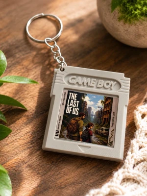 Game Boy nyckelring The Last of Us - Retroinspirerad nyckelring formad som ett Game Boy-spel med motiv från The Last of Us. Tillverkad i grå plast med metallkedja och ring. Perfekt accessoar för gamers och nördar som gillar unika detaljer.