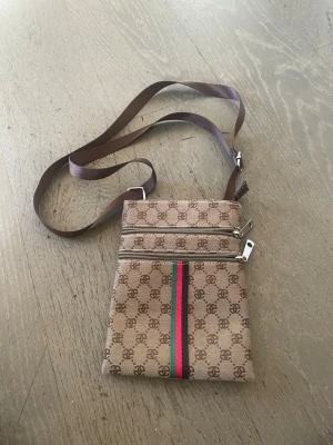 Beige axelväska med monogram och ränder - Snygg beige axelväska med brunt monogrammönster och två dragkedjefack framtill. Väskan har en brun justerbar axelrem och en grön-röd rand som detalj på framsidan. Tillverkad i slitstarkt tygmaterial, perfekt för att bära det viktigaste.