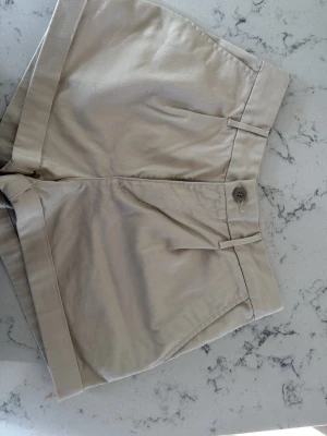 Beige bomullsshorts med knapp levis - Snygga beige shorts i bomull med hög midja och uppvikta benslut. Klassisk design med fickor på sidorna och stängning med knapp och dragkedja framtill. Perfekta för varma dagar och enkel att matcha med olika toppar.