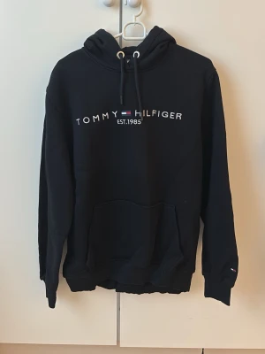 Tommy Hilfiger hoodie - Svart hoodie från Tommy Hilfiger med broderad logga och text. Klassisk känguruficka framtill, justerbar huva med snören och ribbade muddar. Gåva vid köp!