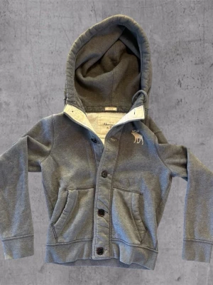 Grå hoodie från Abercrombie & Fitch - Snygg grå hoodie från Abercrombie & Fitch med knäppning framtill och två fickor. Har en broderad älg på bröstet och en mjuk, fodrad huva. Tillverkad i bomullsmix för skön känsla. Perfekt för en avslappnad och trendig look.