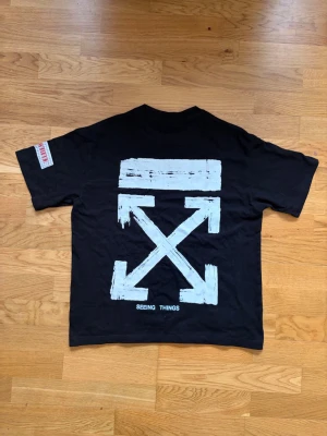 Svart Off-White T-shirt med tryck - Svart t-shirt från Off-White med stort vitt grafiskt tryck på ryggen och texten 'SEEING THINGS' nedtill. Framtill står det 'OFF' och två vita block vid nederkanten. Klassisk rund hals och korta ärmar. Snygg streetwear-vibe. Står storlek M i tröjan men sitter som S/XS